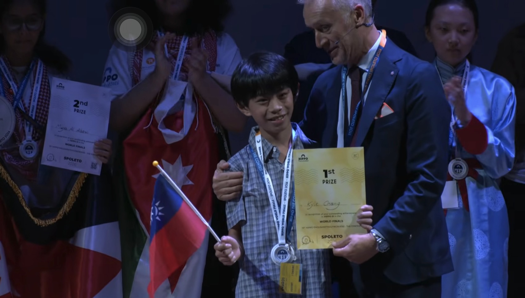Kyle Chang on the World Finals award stage holding his 1st Prize certificate and a Taiwan flag, with the presenter beside him